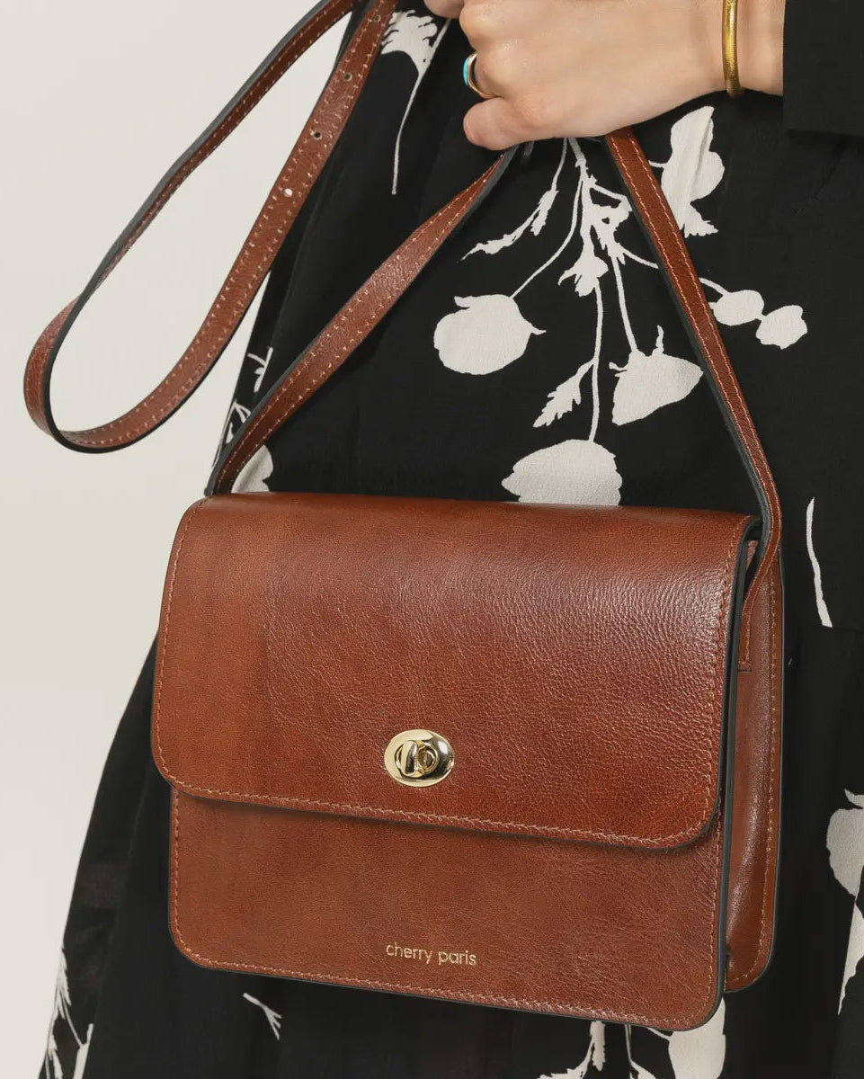 Brown leather handbag with a visible brand logo held by a person wearing a black floral dress.