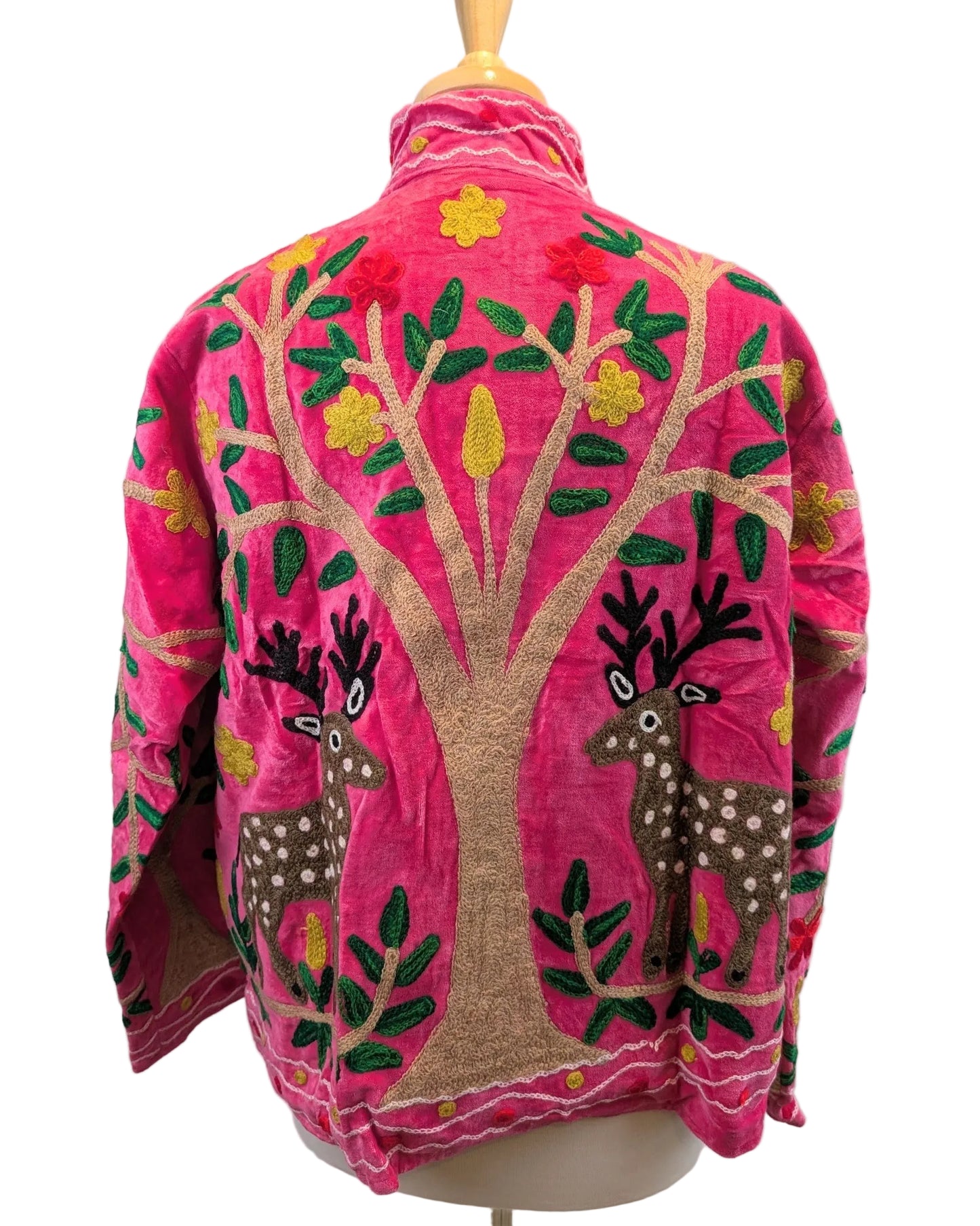A bright pink embroidered jacket displayed on a mannequin, featuring a large tree with green leaves and brown trunks, two deer, and yellow floral accents.