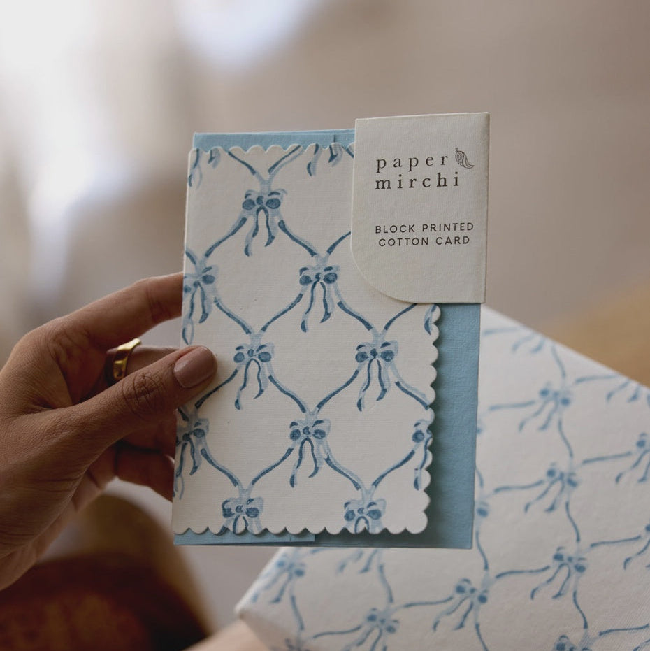 Hand holding a block printed cotton card with blue bow pattern, branded 'paper mirchi'.