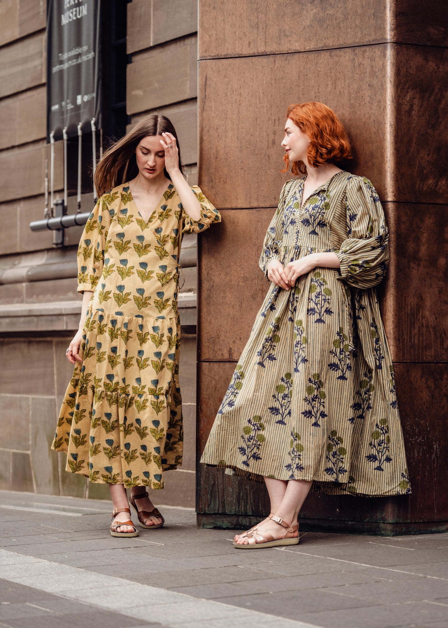 Two women in floral dresses standing against a stone wall.